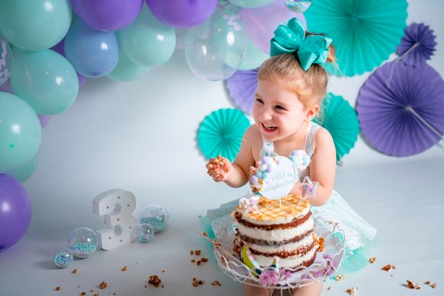 niña pequeña comiendo un pastel de cumpleaños con las manos. de fondo globos azules y morados