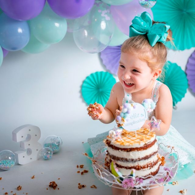 niña pequeña comiendo un pastel de cumpleaños con las manos con globos azules y morados de fondo