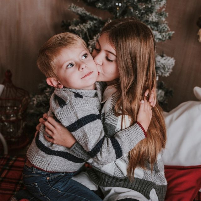 niña dando un beso en la mejilla a su hermano, con decoración navideña de fondo