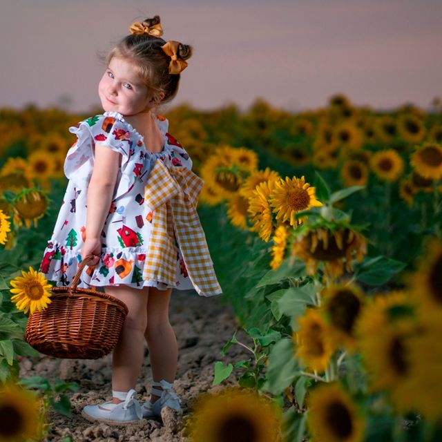 niña con vestido de flores y cesta de mimbre posando en un campo de girasoles