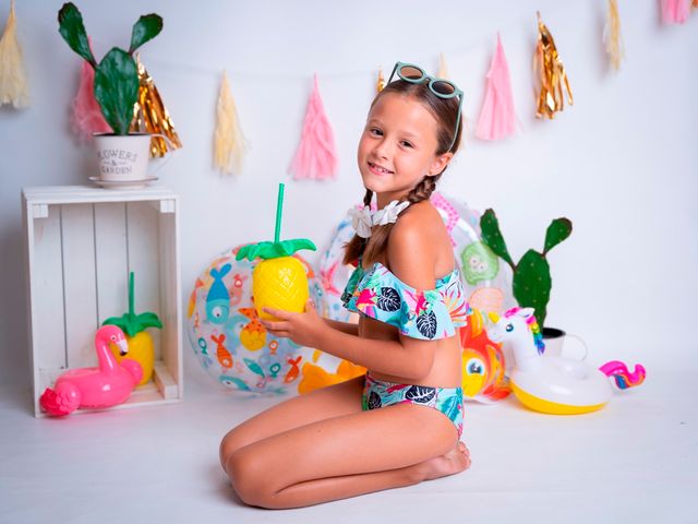 niña de rodillas sosteniendo un vaso en forma de piña, con decoración de playa de fondo