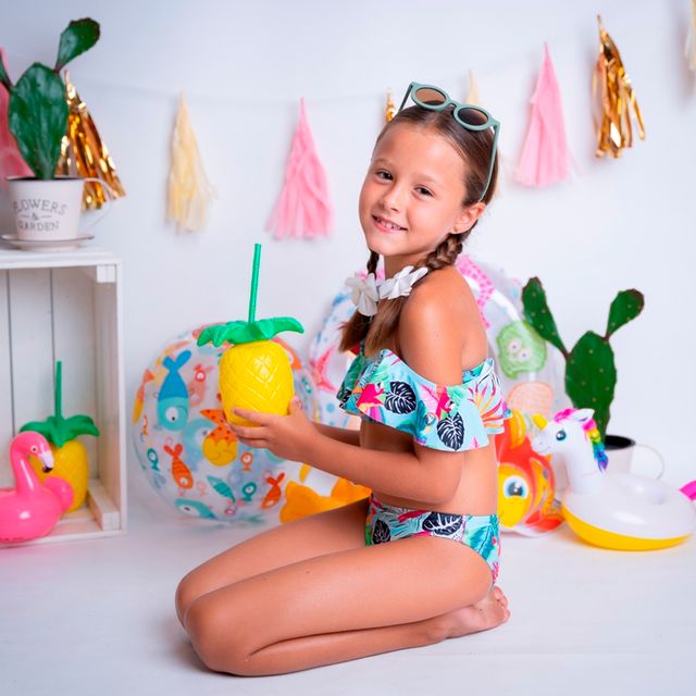 niña de rodillas sosteniendo un vaso en forma de piña, con decoración de playa de fondo