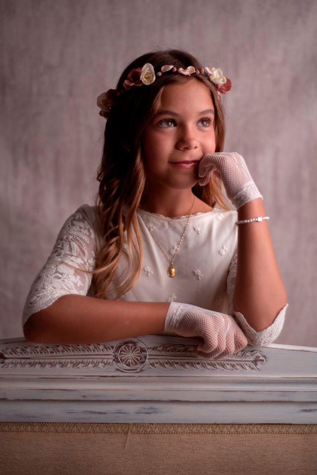 chica en vestido de comunión, con guantes y tiara de flores, apoyada en un mueble