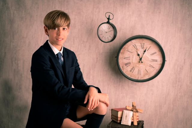 chico en traje de comunión posando sentado junto a un crucifijo, varios libros y relojes colgados en la pared