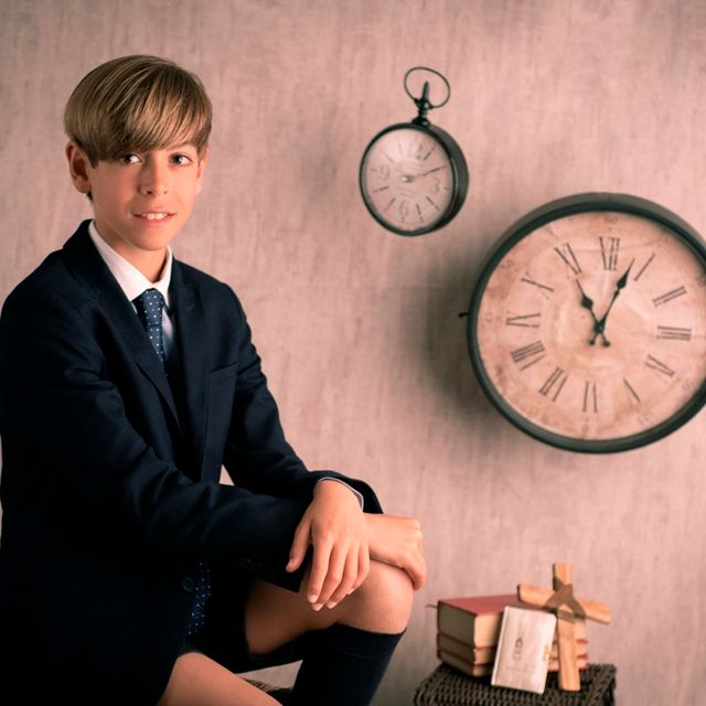 chico en traje de comunión posando sentado junto a un crucifijo, varios libros y relojes colgados en la pared